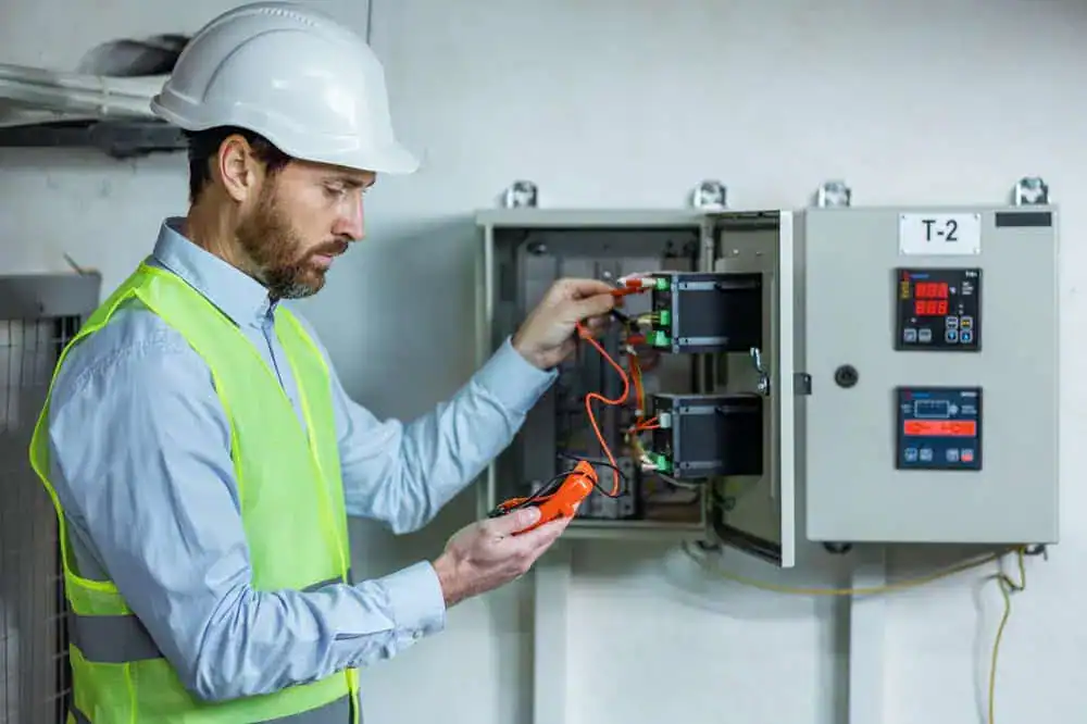 A commercial electrician wearing a white hard hat and reflective vest uses a device to test electrical connections in an open control panel labeled "T-2." They are part of an electrical services team in a work environment focused on safety and precision.