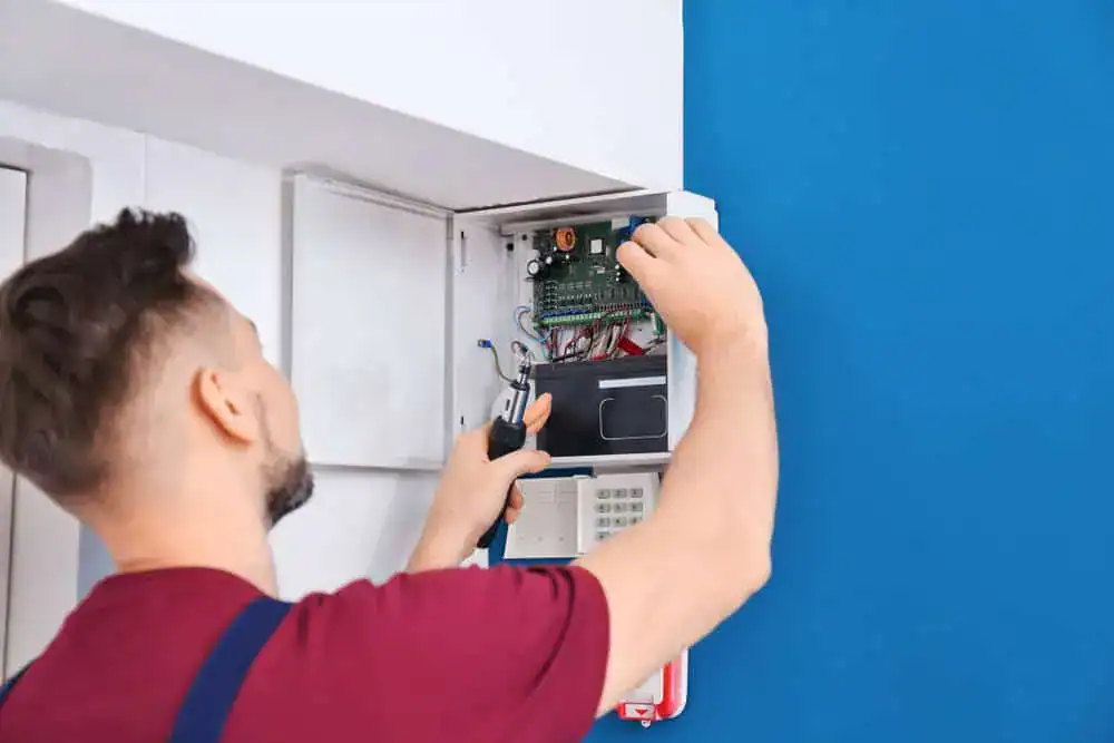 A commercial electrician in a red shirt and overalls skillfully works on wiring within an open electrical panel mounted on a blue wall. He's adjusting components with precision, focusing intently on the connections inside the panel.