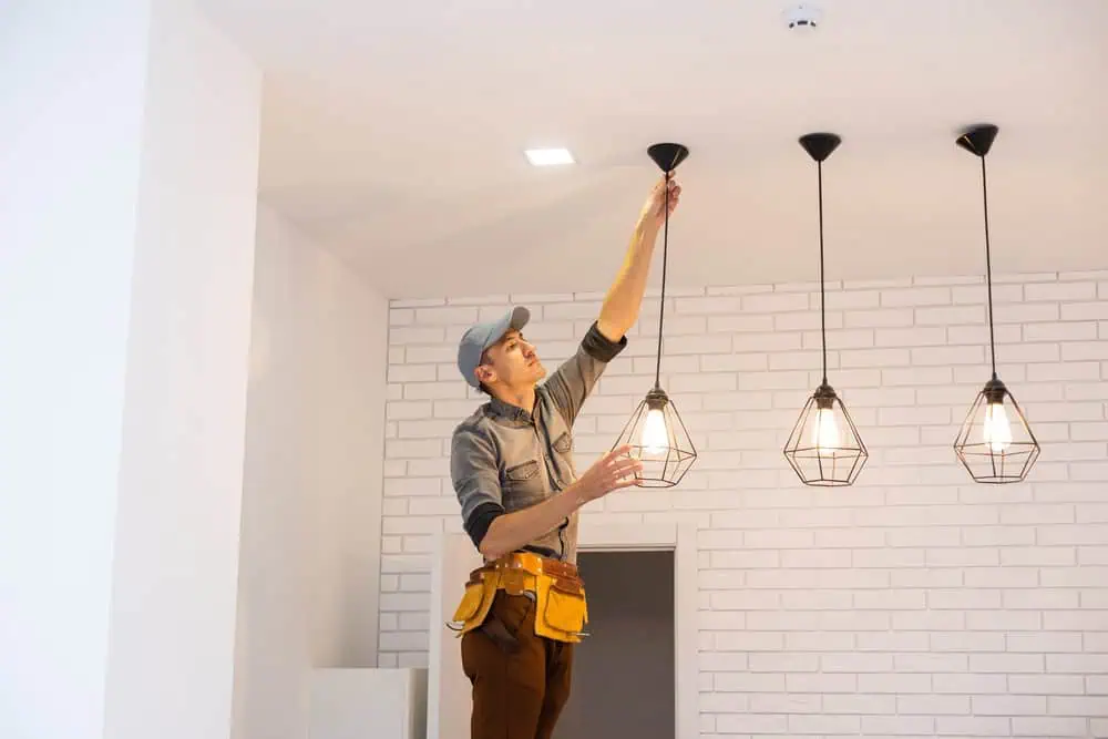 A man in a work uniform and cap stands on a stepladder, adjusting a hanging pendant light fixture in a modern room with white brick walls and three lit lamps.