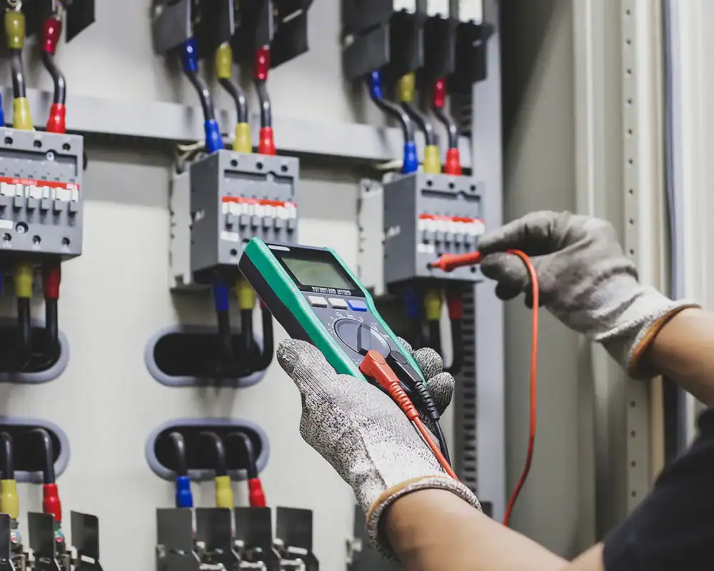 An electrician in Dallas and Fort Worth, TX, wearing protective gloves, uses a digital multimeter to test electrical connections in a control panel with multiple circuit breakers and colored wires.