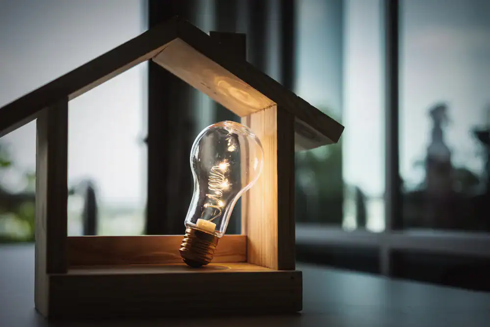 A glowing light bulb sits inside a small wooden house-shaped frame, symbolizing the bright solutions provided by an electrician Dallas and Fort Worth, TX, with blurred windows and outdoor scenery visible in the background.